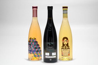 Drei Flaschen Wein stehen nebeneinander: eine rosafarbene, eine schwarze und eine gelbe, jede mit einzigartigem, künstlerischem Etikett.
