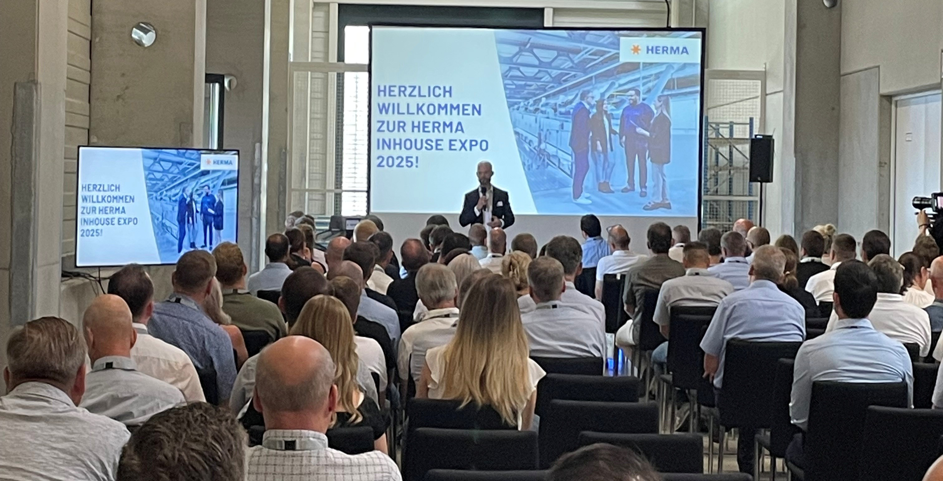 Ein Redner präsentiert auf der HERMA Inhouse Expo 2025 vor einem Publikum. Im Hintergrund ist eine Folie mit dem Willkommensgruß zu sehen.