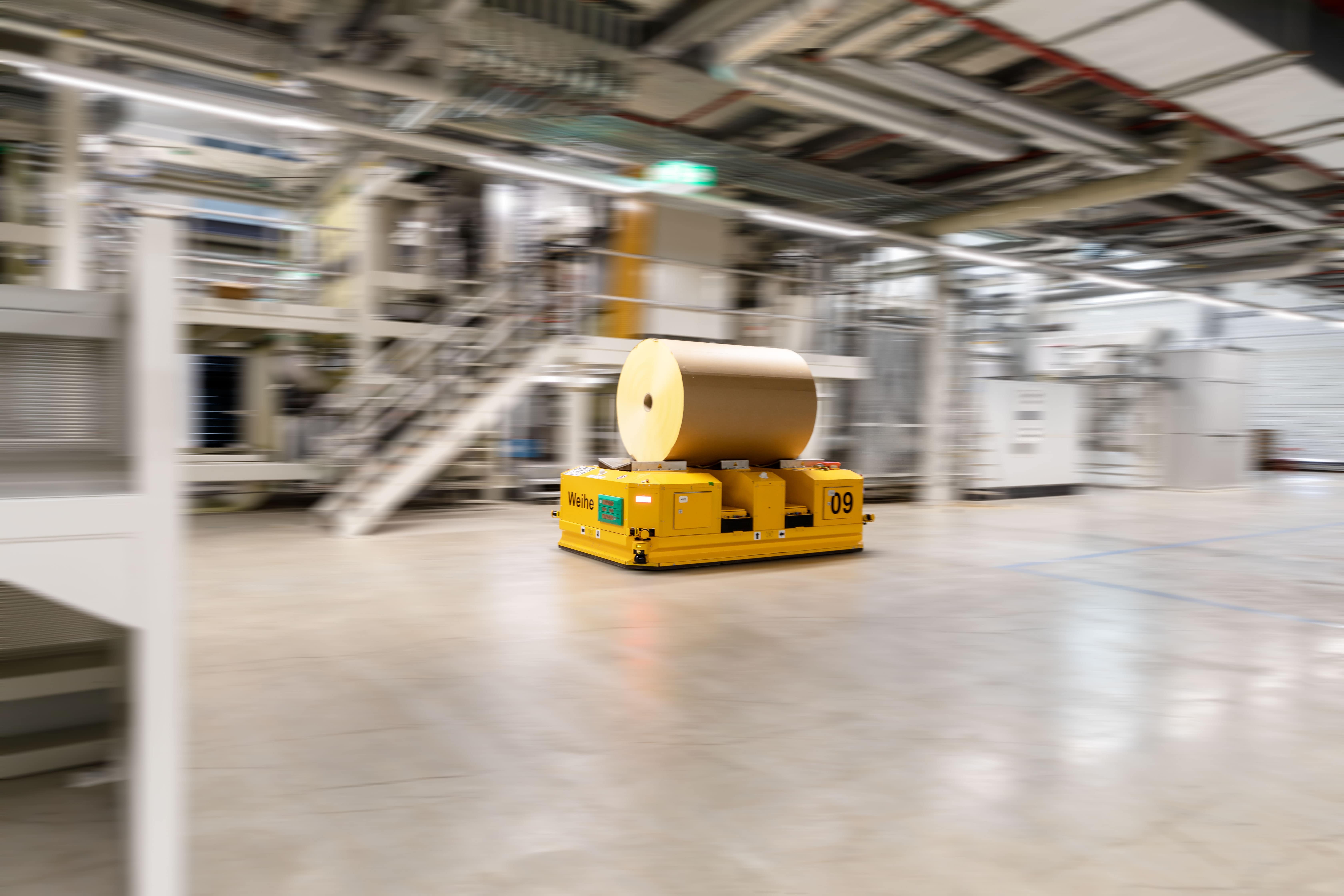 Ein gelber automatisierter Transportwagen bewegt sich mit einer großen Papierrolle in einer modernen Fabrikhalle.
