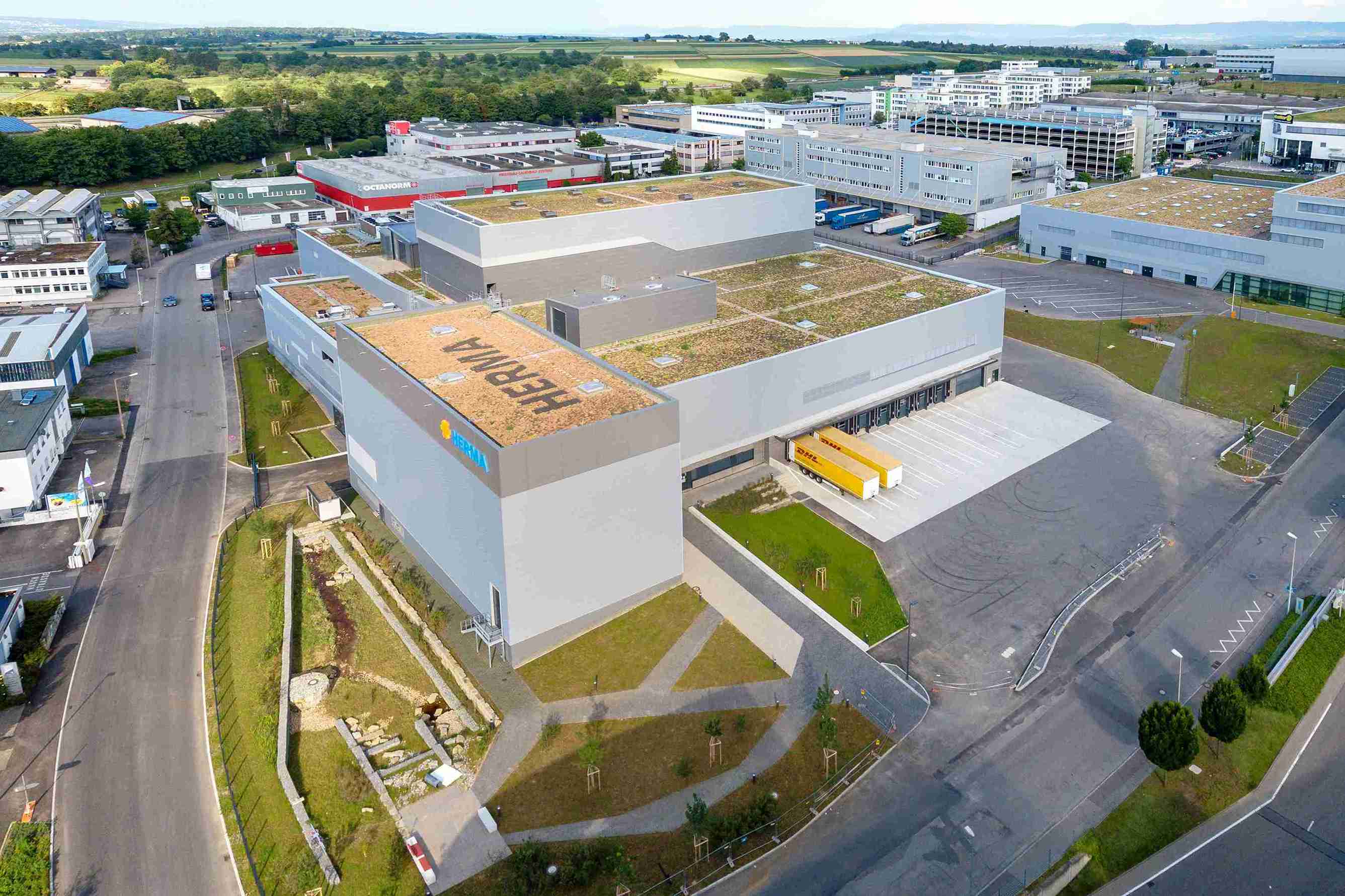 Luftaufnahme eines modernen Industriegebäudes mit begrüntem Dach und umliegenden Parkplätzen, Teil eines größeren Gewerbegebiets.