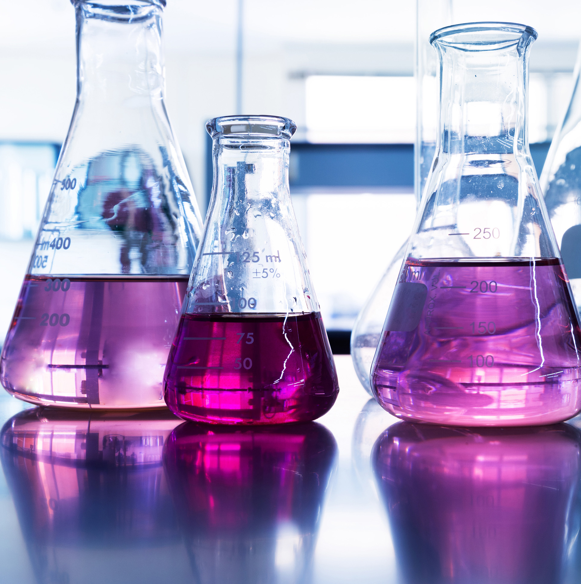 Vier Erlenmeyerkolben mit pinkfarbener Flüssigkeit, beschriftet mit Messskalen, auf einem Tisch im Labor.