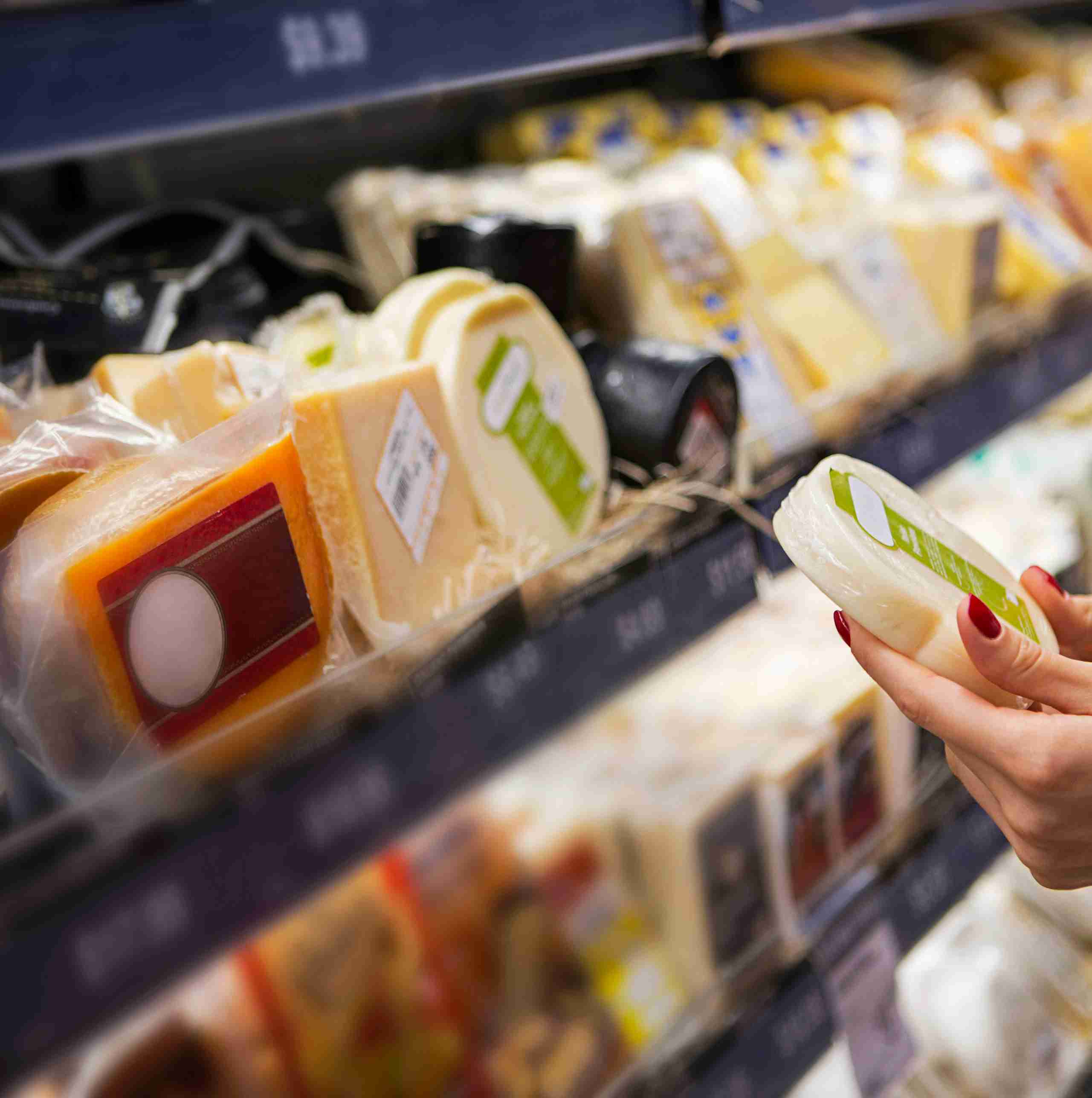 Eine Hand hält ein Stück Käse vor einem Regal mit verschiedenen Käsesorten, die in Verpackungen präsentiert sind.