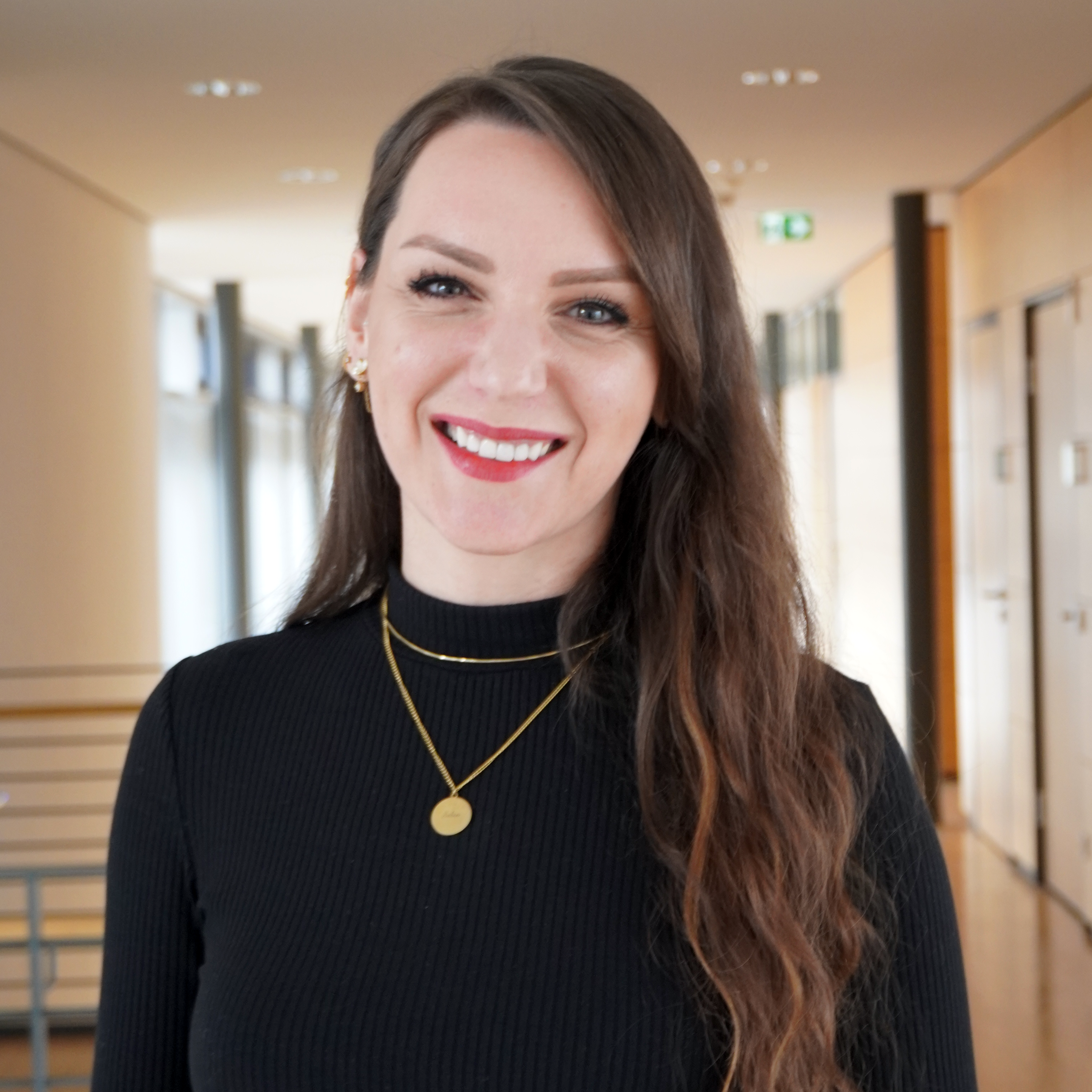 Eine Frau mit langen, lockigen Haaren trägt ein schwarzes Oberteil und Schmuck. Sie lächelt freundlich in einem modernen Innenraum.