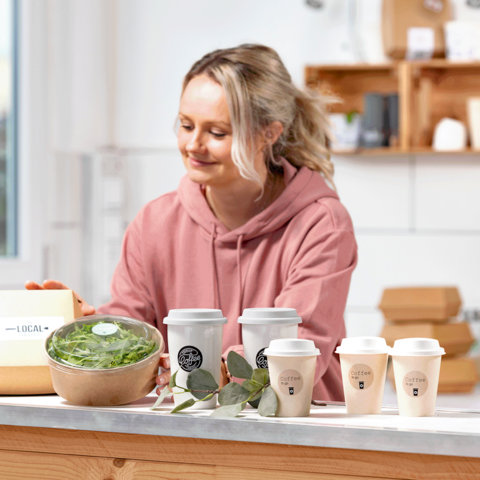 Una mujer con un hoodie rosa está preparando comidas saludables, incluyendo un tazón de ensalada y varias tazas de café sobre una mesa.