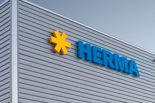 Fassade eines modernen Gebäudes mit dem Logo "HERMA" in blauer Schrift und einem gelben Symbol.