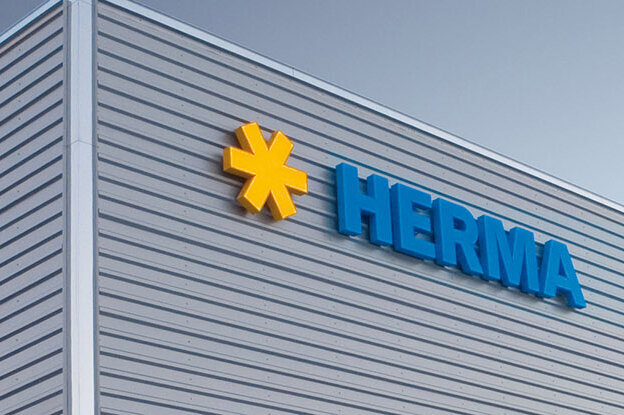Fassade eines modernen Gebäudes mit dem Logo "HERMA" in blauer Schrift und einem gelben Symbol.