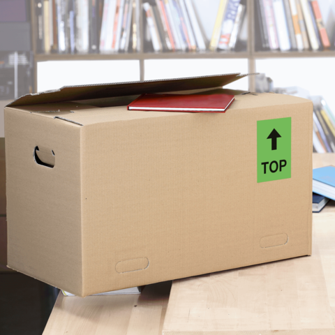 Kartonbox auf einem Tisch mit einem roten Heft und einem grünen "TOP"-Schild. Im Hintergrund sind Bücherregale sichtbar.