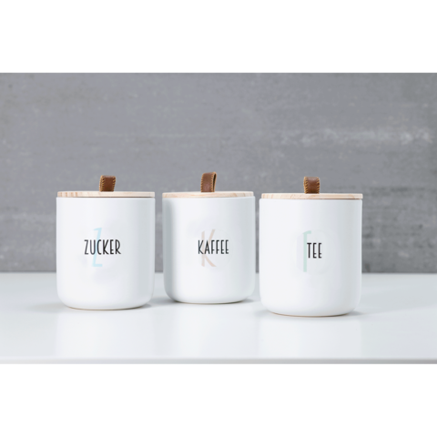 Drei weiße Behälter mit Holzdeckeln, beschriftet mit "Zucker", "Kaffee" und "Tee", auf einem hellen Tisch vor einer grauen Wand.