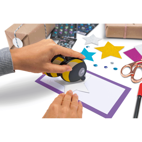 Eine Hand verwendet einen Stempel, um einen Stern auf eine Karte zu drucken. Umgeben von Geschenkverpackungen und Bastelmaterialien.