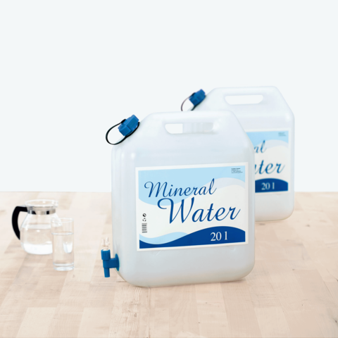 Zwei 20-Liter-Behälter mit Mineralwasser, beschriftet mit "Mineral Water", stehen auf einem Tisch neben einem Wasserglas und einer Karaffe.