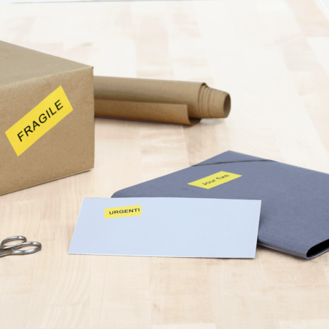 Ein Karton mit dem Aufkleber "FRAGILE", daneben ein graues Kuvert mit "DRINGEND!" und eine Schere auf einem Holztisch.