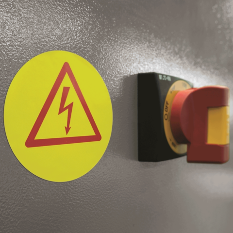 Warnschild mit Blitzsymbol und rotem Not-Aus-Schalter an einer grauen Wand.