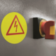 Warnschild mit Blitzsymbol und rotem Not-Aus-Schalter an einer grauen Wand.