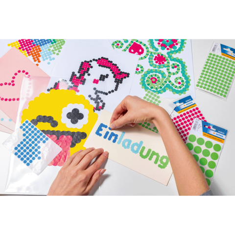 Eine Hand platziert bunte Punkte auf ein Einladungskarte, um ein kreatives Design zu erstellen. Verschiedene Bastelmaterialien liegen auf dem Tisch.