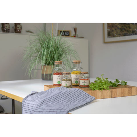 Glasbehälter mit Gewürzen und frischen Kräutern auf einem Holzbrett, daneben ein gestreiftes Küchentuch und eine Pflanze im Hintergrund.