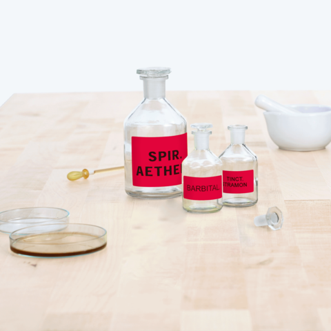 Flaschen mit roten Etiketten, beschriftet mit "Spir. Aether", "Barbital" und "Tinct. Tramon", auf einem Holztisch. Ein Mörser und zwei Petrischalen sind ebenfalls sichtbar.
