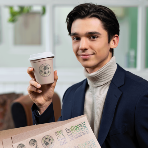Junger Mann in einem Anzug hält einen Kaffeebecher in der Hand und lächelt. Im Hintergrund sind unscharfe Möbel zu sehen.