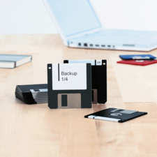 Mehrere Disketten mit Beschriftungen, darunter "Backup 1/4", liegen auf einem Tisch neben einem Laptop.