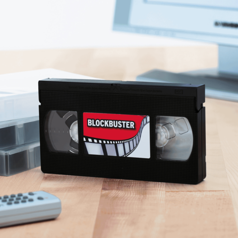 Eine schwarze VHS-Kassette mit einem Blockbuster-Label liegt auf einem Tisch neben einer Fernbedienung und einer leeren Kassette.
