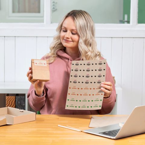 Eine Frau mit blonden, lockigen Haaren hält eine kleine Verpackung und ein Blatt mit Aufklebern in der Hand, während sie an einem Tisch sitzt.