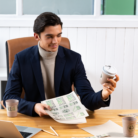 Un joven en traje sostiene una taza de café y una hoja de papel con texto en un entorno de oficina.