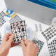 Hände halten eine transparente Box mit Zahlen und Buchstabenaufklebern, umgeben von verschiedenen Aufklebern auf einem Tisch.