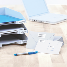 Carpeta con compartimentos etiquetados para documentos entrantes y salientes, junto a sobres y un bolígrafo azul sobre una mesa.