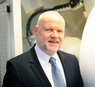 Ein Mann in einem Anzug mit Krawatte lächelt vor einer technischen Maschine. Der Hintergrund zeigt Teile von industrieller Ausrüstung.