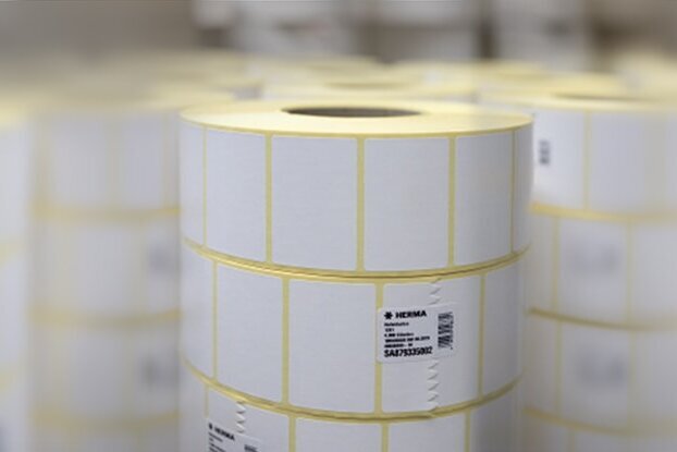 Rollos con etiquetas blancas, apilados en un entorno de almacenamiento. Las etiquetas están marcadas con líneas amarillas.