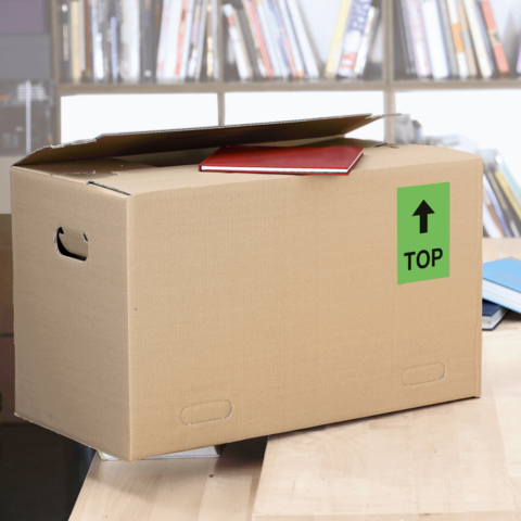 Kartonbox auf einem Tisch, mit einem roten Heft und einem grünen "TOP"-Schild auf der Seite. Hintergrund zeigt Bücherregale.
