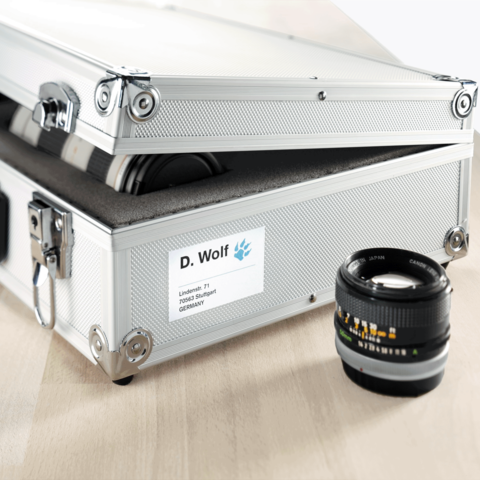 Aluminiumkoffer mit einem Etikett, das den Namen "D. Wolf" zeigt, und einem Kameraobjektiv daneben.