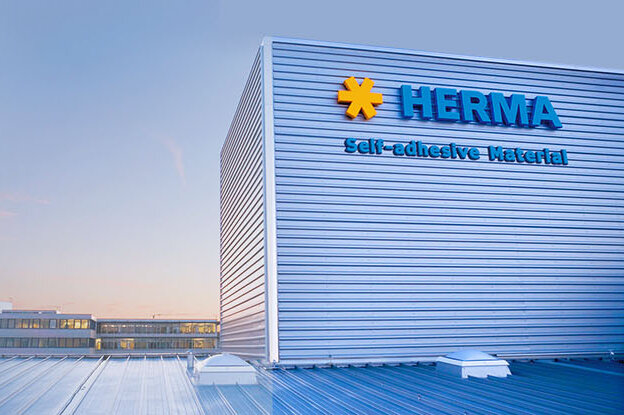 Das Gebäude mit dem Schriftzug "HERMA" und einem blauen Himmel im Hintergrund. Die Architektur ist modern und funktional gestaltet.