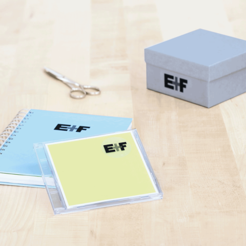 Ein blauer Notizblock, eine gelbe CD-Hülle mit dem Schriftzug "EIF", eine Schere und eine graue Geschenkbox auf einem Holztisch.