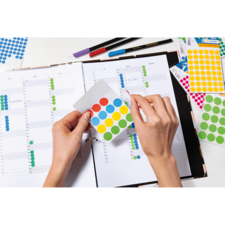 Eine Person platziert bunte Aufkleber auf einem geöffneten Planer, um Termine oder Aufgaben zu kennzeichnen. Stifte liegen daneben.