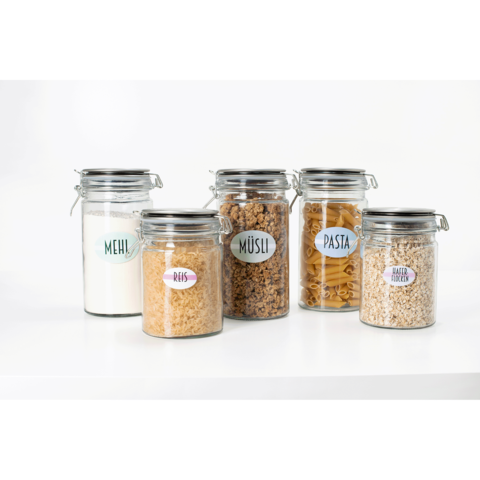 Fünf Glasbehälter mit Etiketten für Mehl, Reis, Müsli, Pasta und Haferflocken, auf einem weißen Tisch angeordnet.
