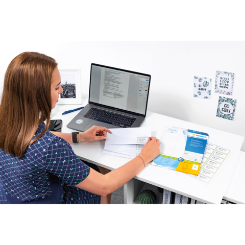 Eine Person sitzt an einem Schreibtisch und arbeitet am Laptop, während sie Dokumente und Briefe auf dem Tisch sortiert.