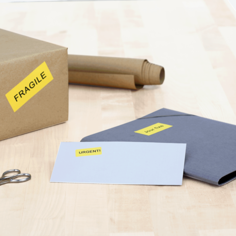 Ein Karton mit dem Aufkleber "FRAGILE", daneben ein graues Kuvert mit der Aufschrift "DRINGEND!" und eine Schere.