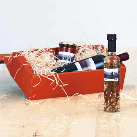 Ein roter Geschenkkorb mit einer Flasche Likör, einem Glas Marmelade und einer Weinflasche, umgeben von Holzschnitzeln.