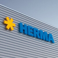 Gelbe Stern-Logos und blaue Buchstaben bilden das Wort "HERMA" an einer modernen Gebäudeaußenwand.