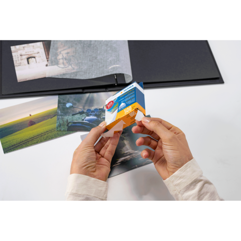 Eine Person hält eine Packung Fotopapier in den Händen, während sie sich auf einen Tisch mit mehreren Fotos und einem Album konzentriert.