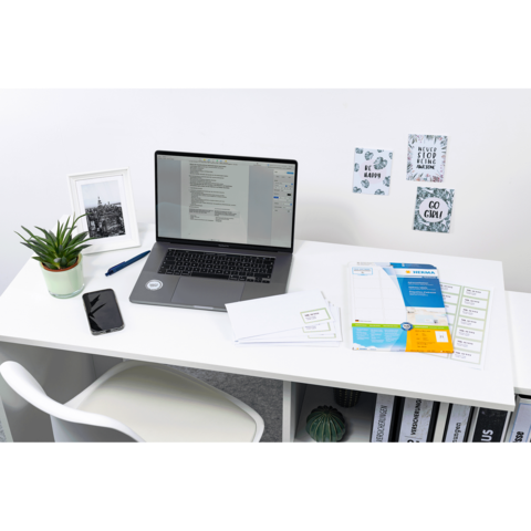 Schreibtisch mit Laptop, Smartphone, Pflanzen und Dokumenten. An der Wand hängen dekorative Notizen und Bilder.
