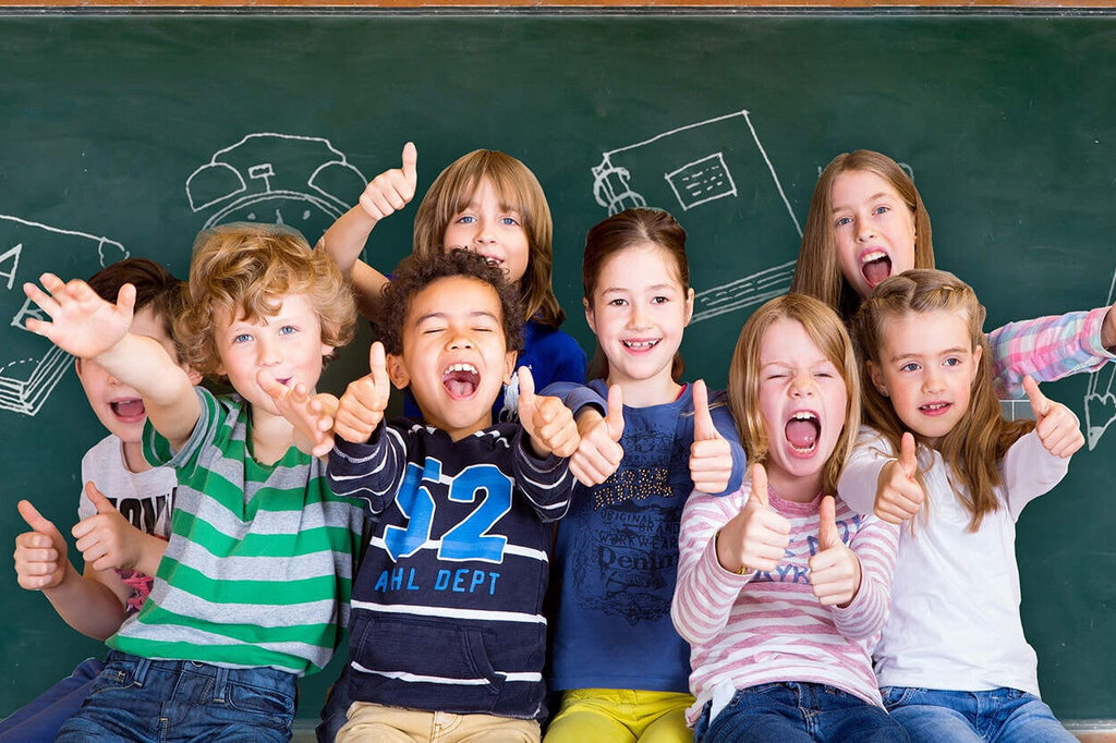 Un grupo de niños está frente a una pizarra, sonriendo y posando alegremente. Detrás se pueden ver dibujos de material escolar.