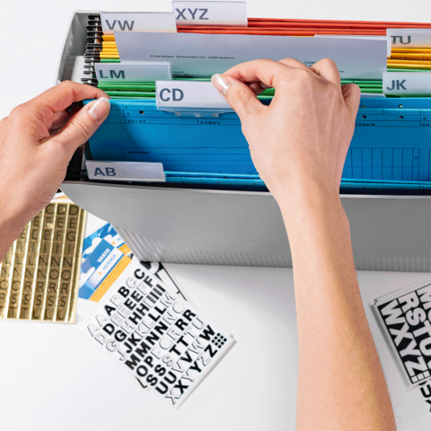 Hände sortieren farbige Ordner in einem Aufbewahrungskasten mit beschrifteten Trennblättern. Daneben liegen Etiketten.