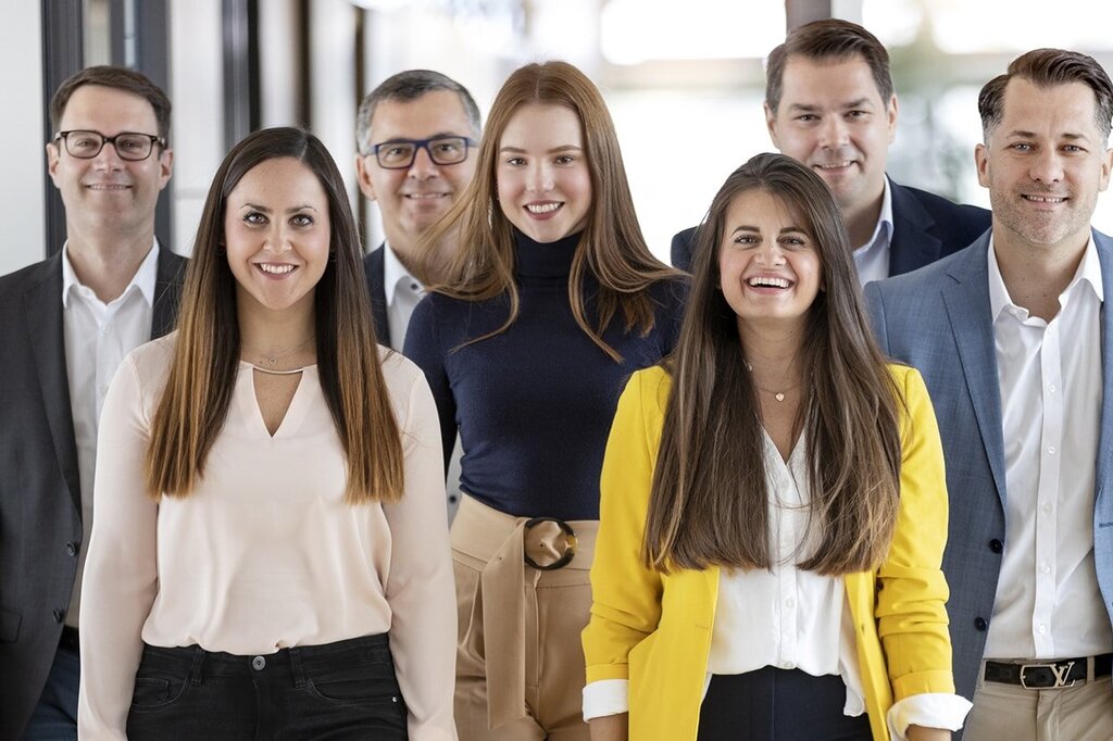 Un grupo de siete personas está en un pasillo, sonriendo y vestidas de manera formal; algunas llevan blusas y trajes.
