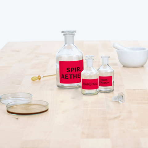 Flaschen mit roten Etiketten, beschriftet mit "Spir. Aether", "Barbital" und "Tinct. Tramon", sowie ein Mörser und zwei Deckgläser auf einem Tisch.