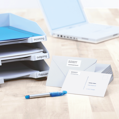 Ordner mit beschrifteten Fächern für eingehende und ausgehende Dokumente, daneben Umschläge und ein blauer Stift auf einem Tisch.