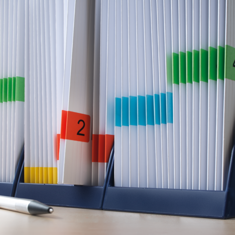 Carpeta con etiquetas de colores y documentos en un soporte, al lado un bolígrafo.