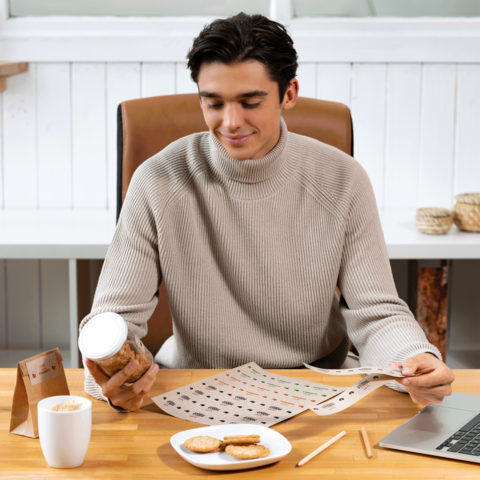 Ein junger Mann sitzt an einem Tisch, betrachtet ein Blatt mit Mustern und hält ein Glas mit Keksen in der Hand. Vor ihm stehen ein Laptop und eine Tasse Kaffee.