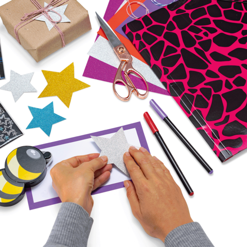 Manos que pegan una estrella brillante en un papel colorido, rodeadas de materiales de manualidades como tijeras, lápices y papel de regalo.