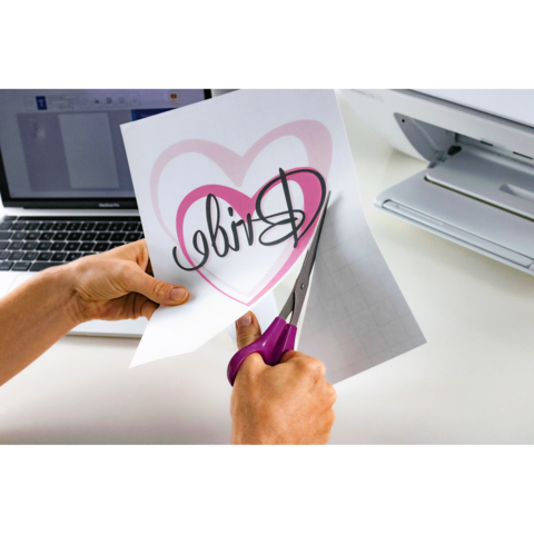 Eine Person schneidet mit einer Schere ein Blatt Papier mit dem Wort "Bride" und einem Herzmuster aus, während ein Laptop im Hintergrund sichtbar ist.